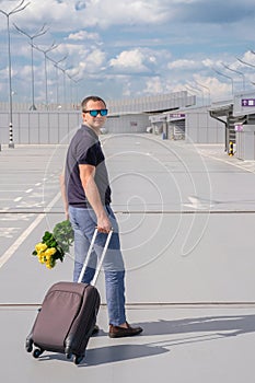 A man with a suitcase and flowers in an airport car park. Business trip. Vertical image