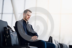 Man with suitcase sitting in airport waiting area while listening music using airpods