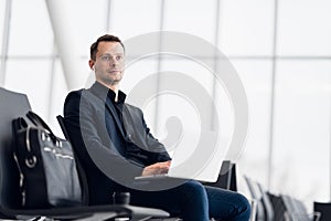 Man with suitcase sitting in airport waiting area while listening music using airpods