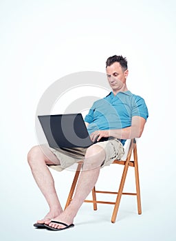 Man in summer clothes is sitting on a chair, typing on a laptop. Light background. Concept of work on vacation.