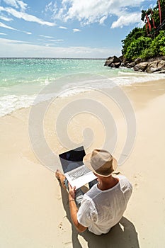 Man in the summer hat , a businessman, digital nomad working with laptop on the sandy beach. Freelancer. Tropics