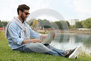 Man in sunglasses with laptop on green grass near lake