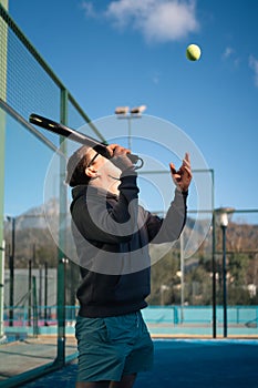 A man in sunglasses serves a tennis ball, enjoying a paddle tennis match on a sunny outdoor court with mountains in the