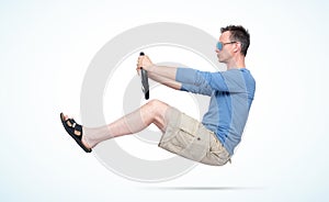 Man in sunglasses, shorts, blue t-shirt and sandals drives car with a steering wheel, on light day background. Auto driver concept