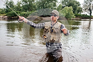 Man in sunglasses stands in water and fishing. He is preparing fly-fishing. Guy holds it in one hand and spoon in the