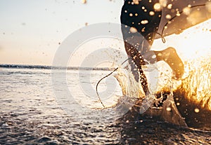 Man surfer run in ocean with surfboard. Closeup image water splashes and legs, sunset light