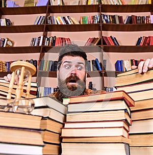 Man on surprised face between piles of books, while studying in library, bookshelves on background. Teacher or student