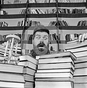 Man on surprised face between piles of books, while studying in library, bookshelves on background. Teacher or student