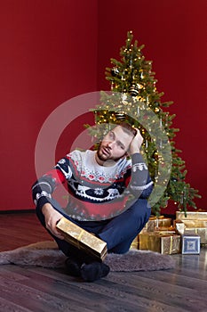 Man in sweater sits by Christmas tree and thinks about gift