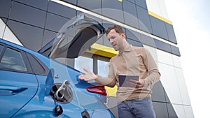 Man with tablet device waiting till his electric car charge. Male has negative emotions, something is not working