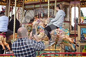 Man taking a photo on a mobile phone of teenager riding on a carousel horse on fairground ride at amusement fair