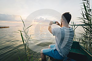Man taking picture on his phone of sunrise while sitting in old boat