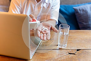 Man taking pill with glass of water and laptop on wooden table.