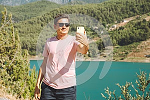 Man taking selfie on mobile phone of mountains lake background. Traveler male having video chat on cellphone on the blue