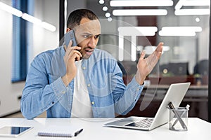 A man is talking on his cell phone while sitting at a desk with a laptop