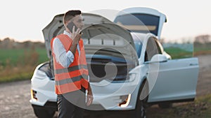 Man talking on the phone and looking at the car`s engine. young man use smartphone stading on road near the broken car