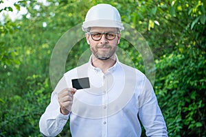 Man taskmaster in hardhat showing blank contact card outdoors, copy space