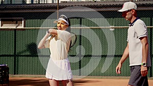 A man tennis coach comes closer her ward and shows how to hold racket properly on shoulder to repel a blow shot of rival