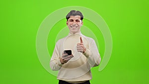 A man is texting on a smartphone. He is looking up and smiling. He is pointing his finger up. He is standing on a green