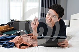 Man thinking with tablet and packing clothes into suitcase on a bed at home, planning travel holiday