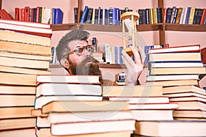 Man on thoughtful face holds hourglass while studying, bookshelves on background. Man, scientist in glasses looks at