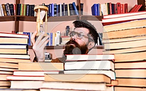 Man on thoughtful face holds hourglass while studying, bookshelves on background. Man, scientist in glasses looks at