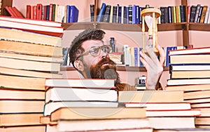 Man on thoughtful face holds hourglass while studying, bookshelves on background. Man, scientist in glasses looks at