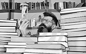 Man on thoughtful face holds hourglass while studying, bookshelves on background. Man, scientist in glasses looks at