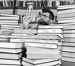 Man on thoughtful face holds hourglass while studying, bookshelves on background. Teacher or student with beard studying