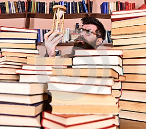 Man on thoughtful face holds hourglass while studying, bookshelves on background. Teacher or student with beard studying