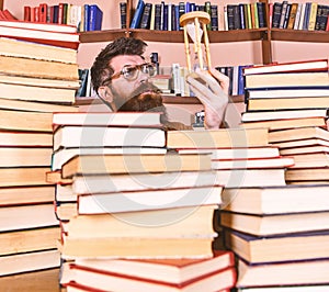 Man on thoughtful face holds hourglass while studying, bookshelves on background. Teacher or student with beard studying