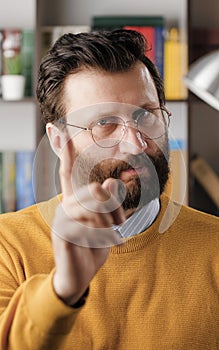 Man threatens with his finger. Serious frowning bearded man in glasses in office or apartment room looking at camera and