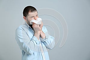 Man with tissue suffering from runny nose on light grey background. Space for text