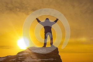 Man at the top of the mountain, holding up his hand, against the background of a dramatic sky in the sunset.