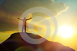 A man at the top of the mountain, raising his hands up against the backdrop of the glare of the sun.