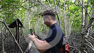 Man Tourist with Backpack Makes Video on Smartphone in Green Mangroves Forest