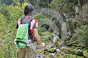 Man tourist in bright clothes with a stick standing on the mountain, looking at the waterfall and the stone mountains