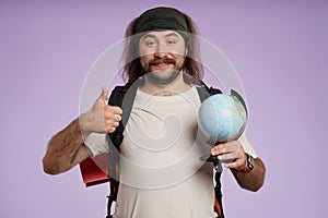 Man tourist in casual clothing with backpack holding globe and shows thumbs up. Isolated on purple background. Portrait