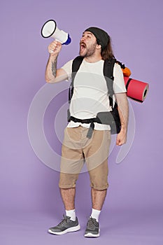 Man tourist in casual clothing with backpack holding megaphone and screaming in it. Isolated on purple background