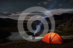 Man tourist with flashlight near his camp tent at night.