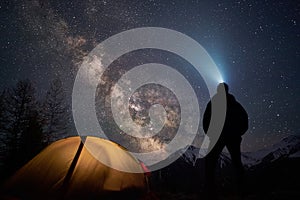 Man tourist with flashlight stands near his camp tent at night under a sky full of stars.