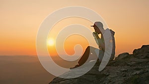 Man tourist sits sitting on edge of cliff, drinks tea from mug, enjoying sunset