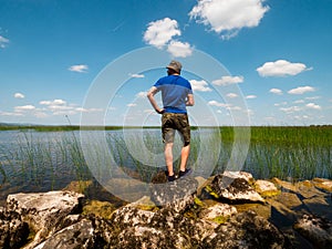 Man tourist standing on a stones by a big lake on a warm sunny day. Blue cloudy sky in the background. Travel to explore nature on