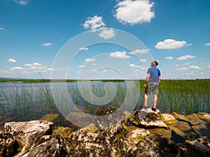 Man tourist standing on a stones by a big lake on a warm sunny day. Blue cloudy sky in the background. Travel to explore nature on