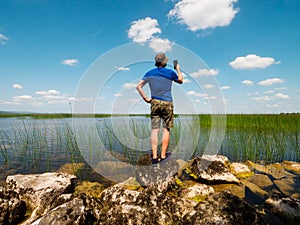 Man tourist standing on a stones by a big lake on a warm sunny day and taking pictures on phone. Blue cloudy sky in the background