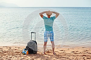 Man tourist in summer clothes with a suitcase in his hand, looking at the sea on the beach, concept of time to travel