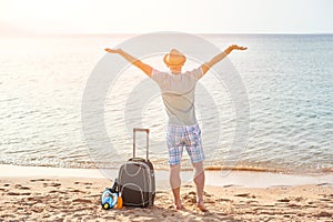 Man tourist in summer clothes with a suitcase in his hand, looking at the sea on the beach, concept of time to travel