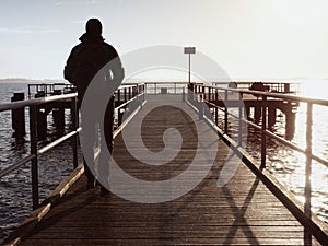 Man tourist at wooden sea pier. Cold windless morning with gradient sunlight. Smooth water