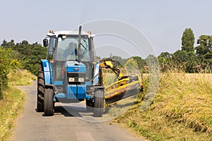 Man in a tractor using a flail to cut grass verges in a country lane. UK