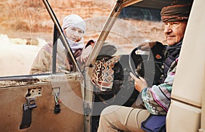 Man in traditional Bedouin coat - bisht - and headscarf, posing behind wheel in old 4wd vehicle, looking to side, younger woman photo